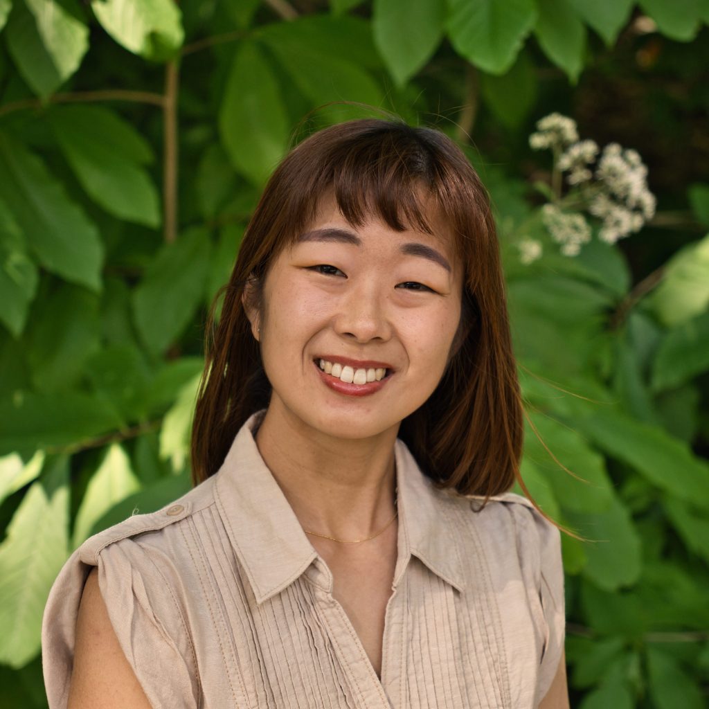 講師プロフィール photo of the instructor Nagi sensei smiling outside in front of green leaves